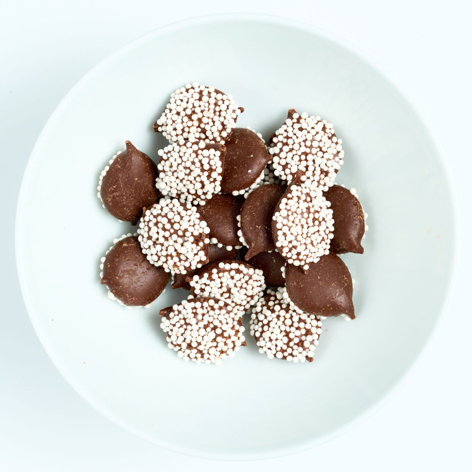 Dark chocolate nonpareils topped with tiny white sprinkles, round candies arranged on a white plate.